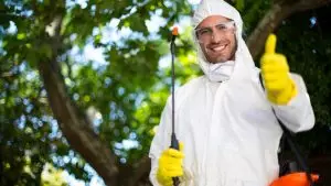 A smiling pest control technician dressed in protective gear and holding a spray nozzle