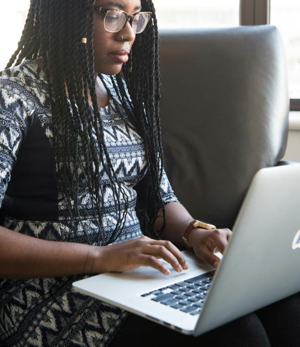 A person with braided hair is sitting on a sofa, focused on typing on a laptop. They are wearing glasses and a patterned dress. Sunlight filters through a nearby window, creating a calm atmosphere.