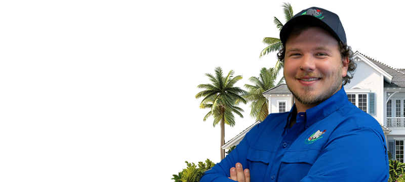 Smiling man in a blue shirt and cap stands with arms crossed in front of a large white house surrounded by palm trees.