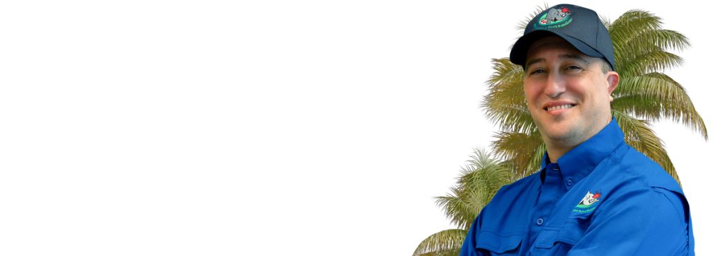 A person wearing a blue shirt and a cap smiles in front of a palm tree. The shirt has an embroidered logo. The background is mostly white, with the person positioned to the right.