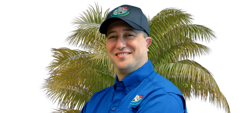 A person wearing a blue shirt and a dark cap with a logo, standing in front of a palm tree, smiling. The background is white.