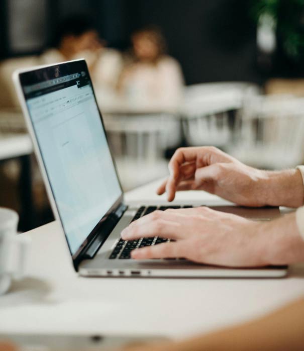 Close-up of hands typing on a laptop keyboard in a dimly lit room. The screen displays a blurred document or webpage. In the background, out of focus, are people sitting at tables.